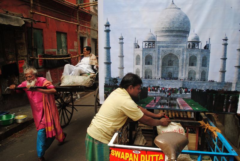 A man pulling a hand rickshaw with a passenger past another man leaning over a cart, in front of a large backdrop printed with the Taj Mahal