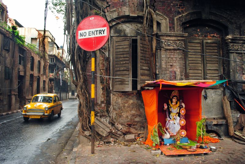 A yellow car on a wet street passing a red No Entry sign beside a small orange-draped roadside shrine with a garlanded statue, in front of a weathered shuttered building