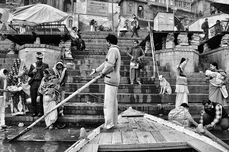 A man standing at the bow of a wooden boat holding a long pole, with people and a dog on the stone steps behind him, in black and white