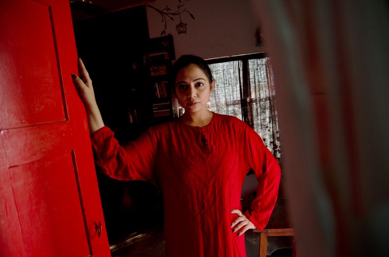 A woman in a long red tunic stands in a dim room beside an open red door, one hand raised against it and the other on her hip, looking into the camera, with bookshelves and a curtained window behind her