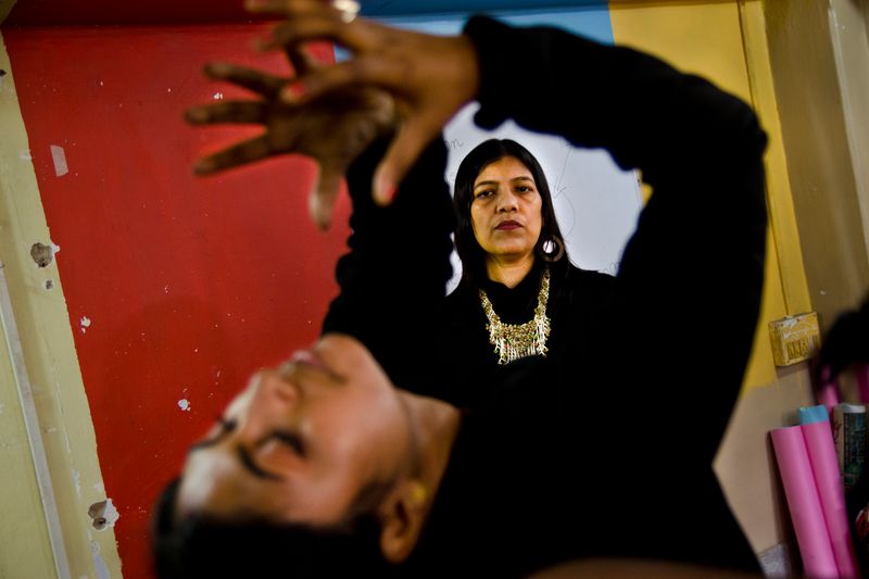 A woman in black with a gold necklace and hoop earrings looks into the camera, framed by the raised arms and spread hands of a blurred figure leaning back in the foreground, against red and yellow painted walls