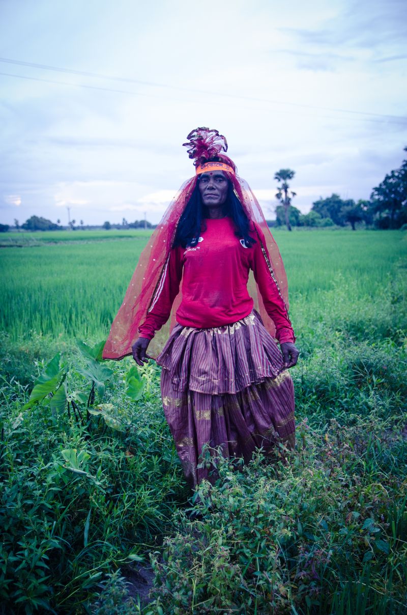A person with long dark hair in a red top, striped skirt, sheer red veil and red ruffled headpiece standing in a green field under a pale cloudy sky