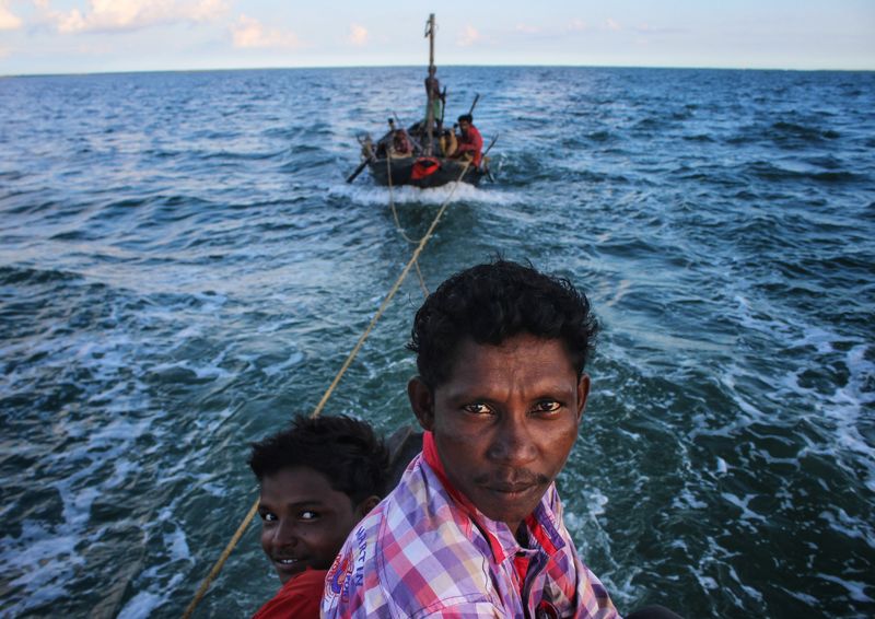 A man and a child at the edge of a boat on open blue water, with two taut ropes leading to another boat with several people aboard behind them