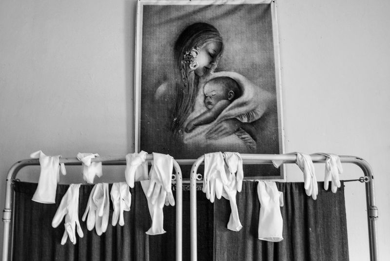 White gloves draped over the top rails of a metal curtain frame, beneath a framed picture of a long-haired person cradling a sleeping baby, in black and white