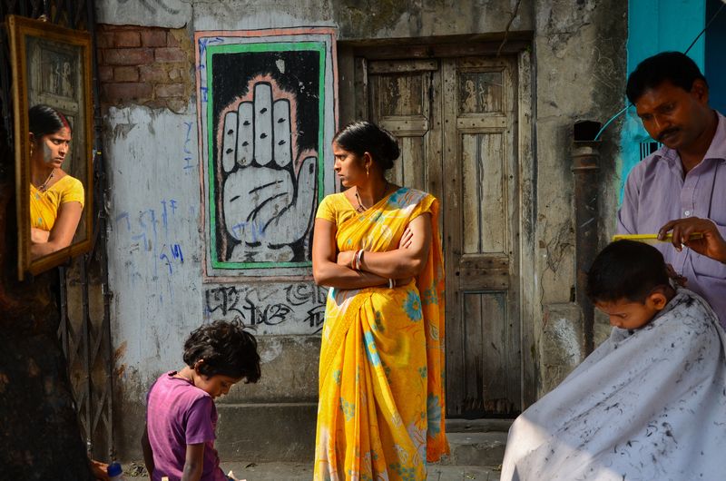 A man combing a cloth-draped child’s hair as a woman in a yellow sari stands with folded arms beside a painted hand mural, with a mirror and a crouching child nearby