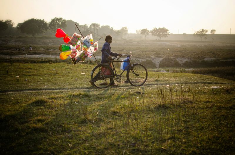 A man pushing a bicycle with a tall pole of colourful balloons and inflatable toys across a grassy field in hazy low light, with trees on the horizon