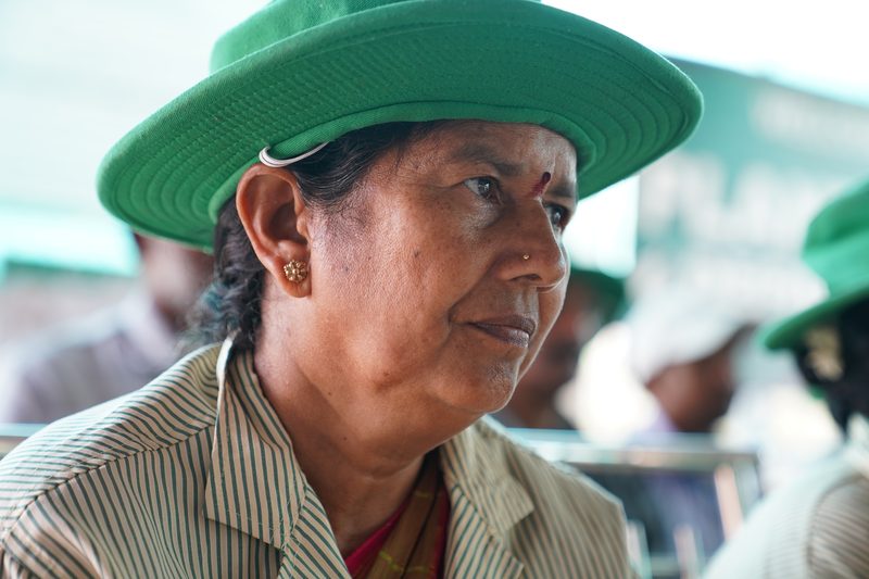 Close profile of a woman in a green brimmed hat and striped collared shirt, her expression calm, with figures blurred in the background.