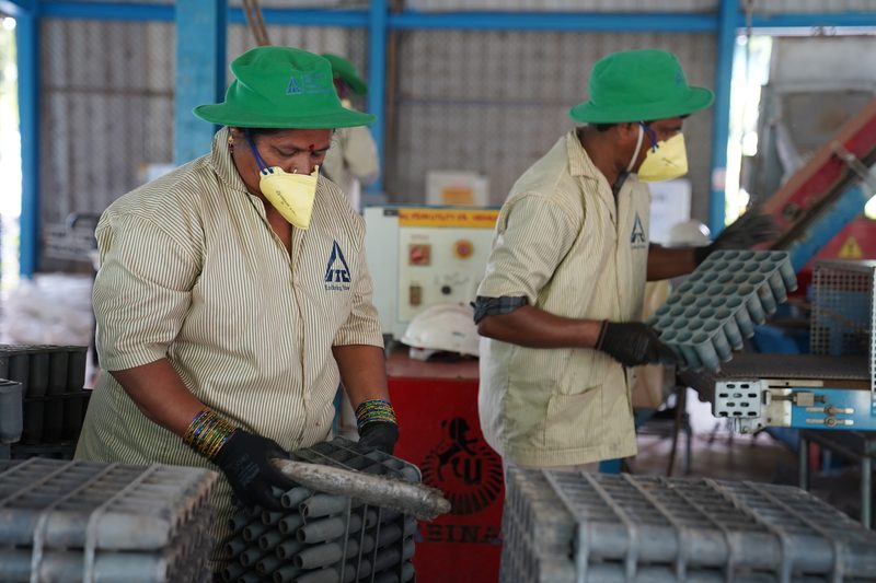 Two people in green hats, yellow face masks and black gloves lift stacks of grey tubular trays in a shed with blue steel pillars.